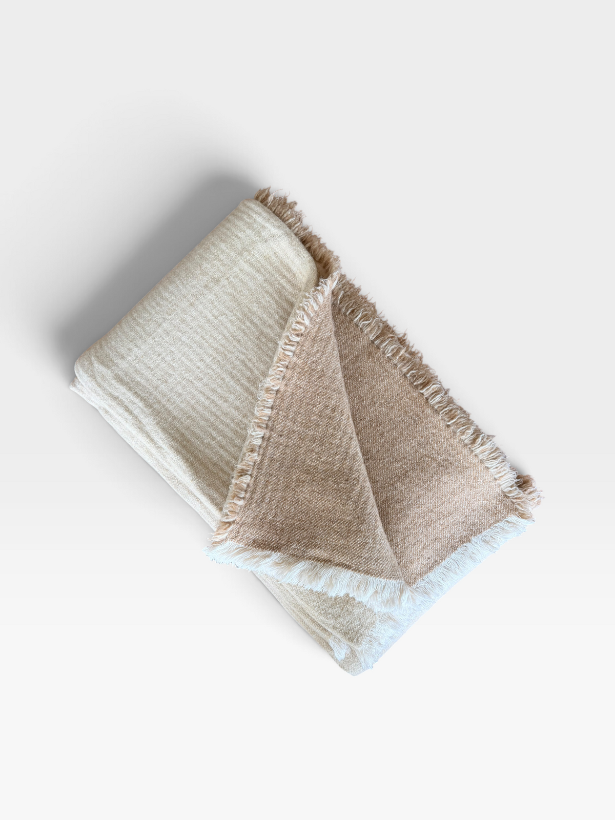 A folded woven throw blanket with fringed edges, shown on a clean white background. The blanket has a textured cream outer side and a softer tan inner side.
