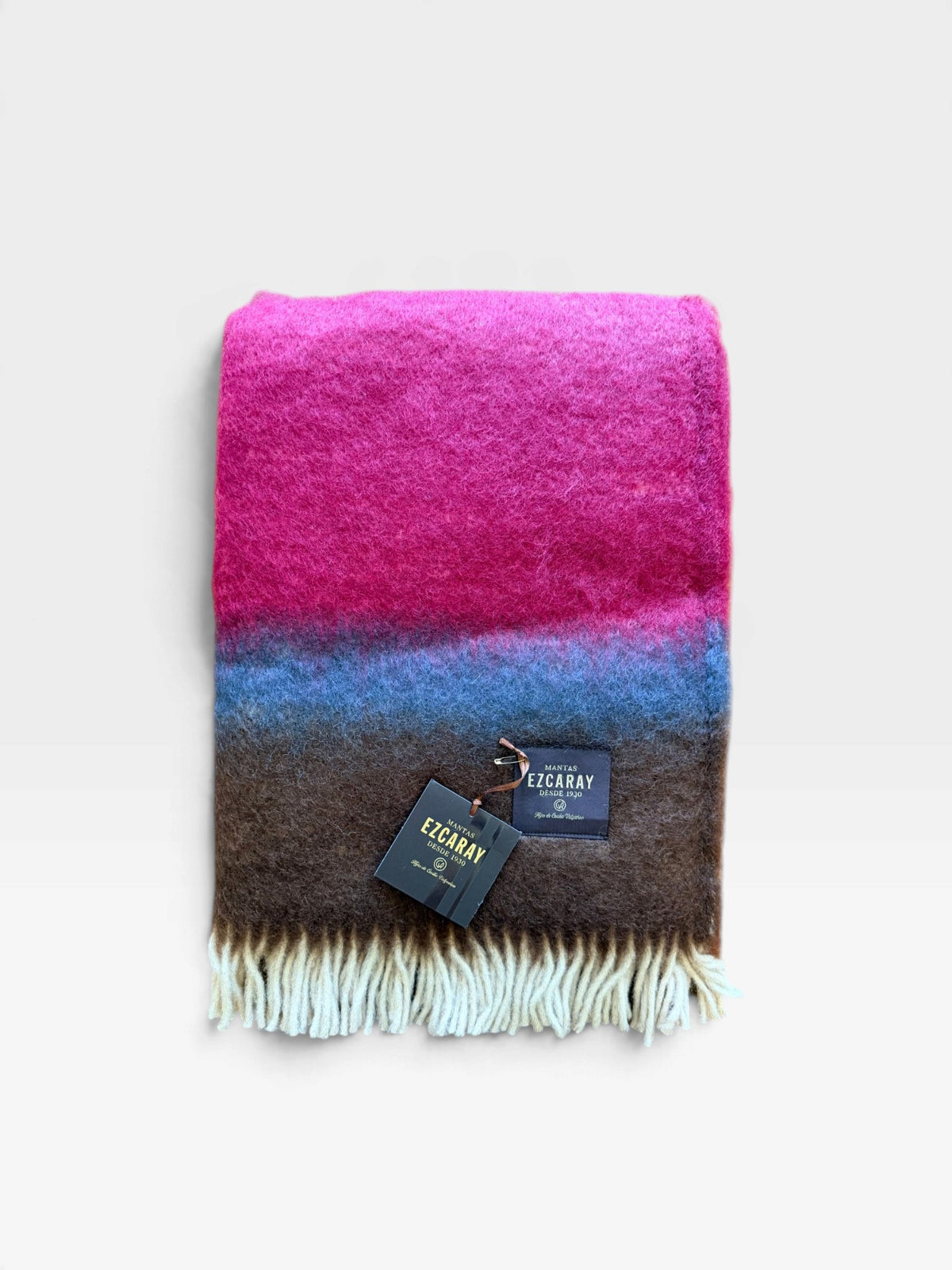 A fringed mohair blanket in hues of magenta, blue, and deep brown folded and sitting on a white background