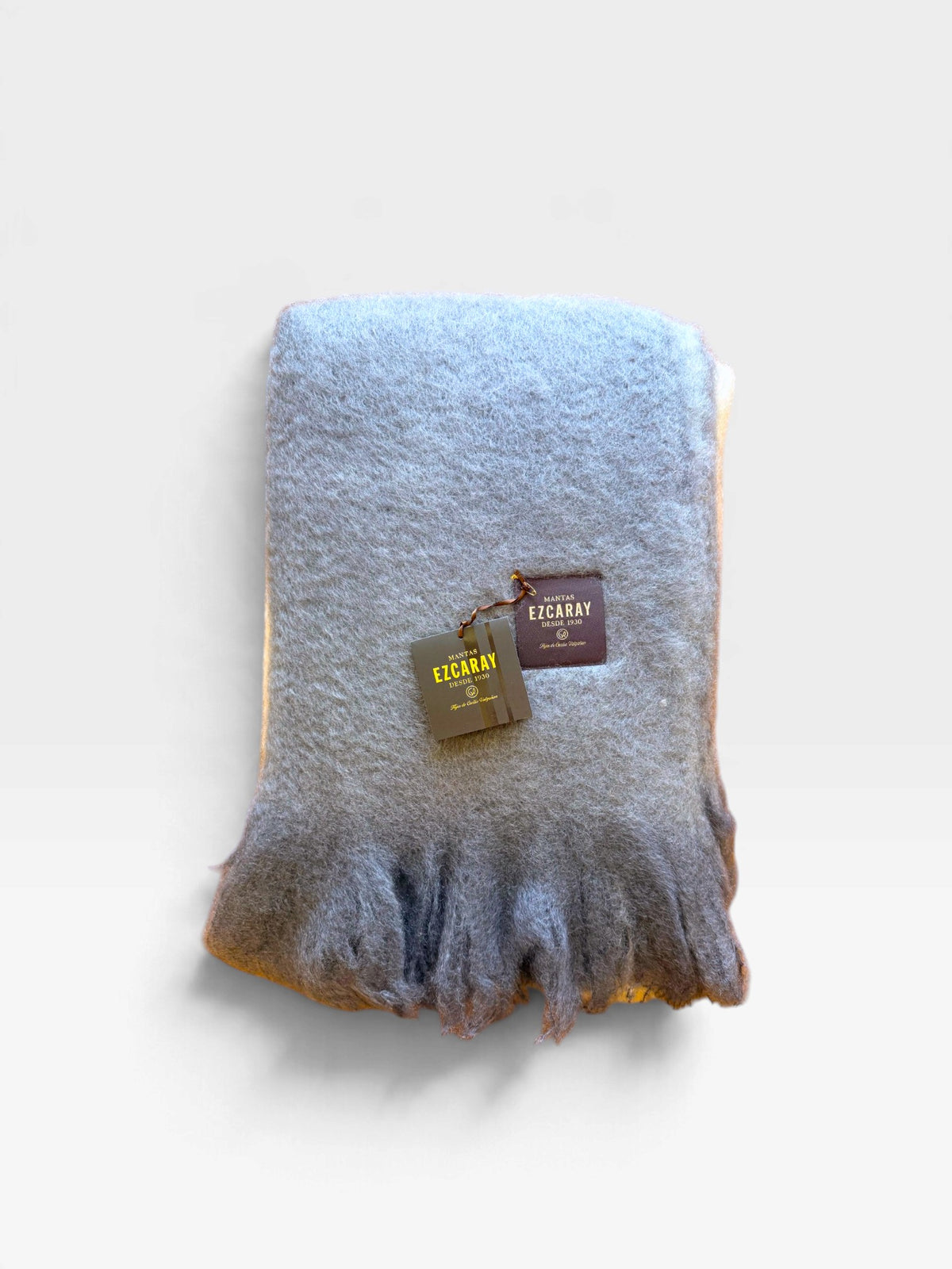 A Mantas Ezcaray mohair throw blanket in shades of gray, ivory, and ochre folded and sitting on a white background