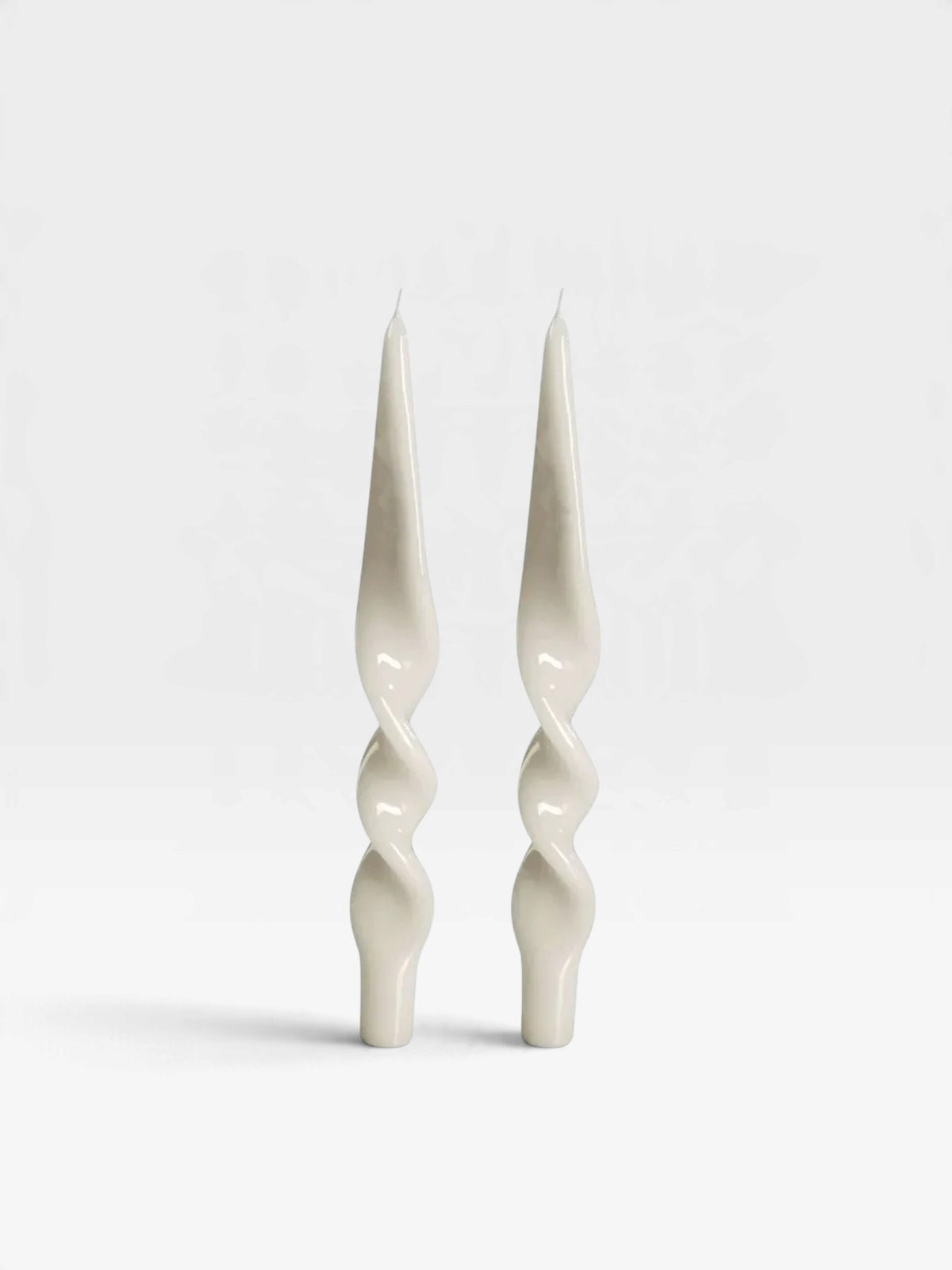 A pair of two lacquered white candles in a twisted design set against a white backdrop