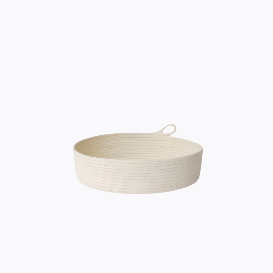 A small, round, beige rope basket with a handle, made from natural cotton, against a white background.