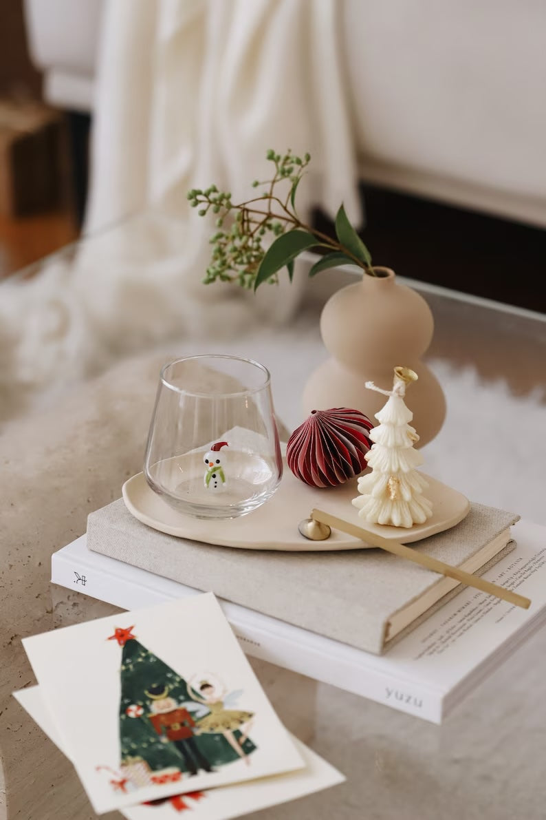 A clear glass tumbler with a colorful snowman figurine perched at the bottom of the glass placed within a Christmas-themed tabletop display.