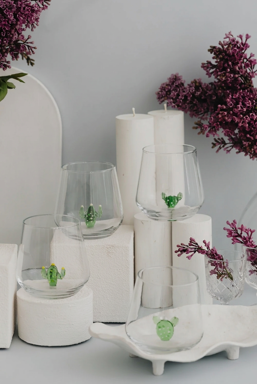 Four glass tumblers with glass cactus-shaped figurines perched at the bottom arranged on a set of white pedastals of differing heights with some lilacs in the background and a gray backdrop