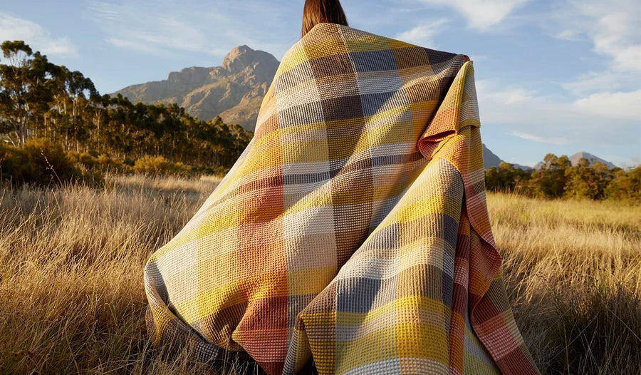 A woman standing in a field with her back to the camera and facing a mountain range in the distance and wrapped in a woven cotton blanket with a plaid-like design in yellow, gray, orange, and white, and beige.
