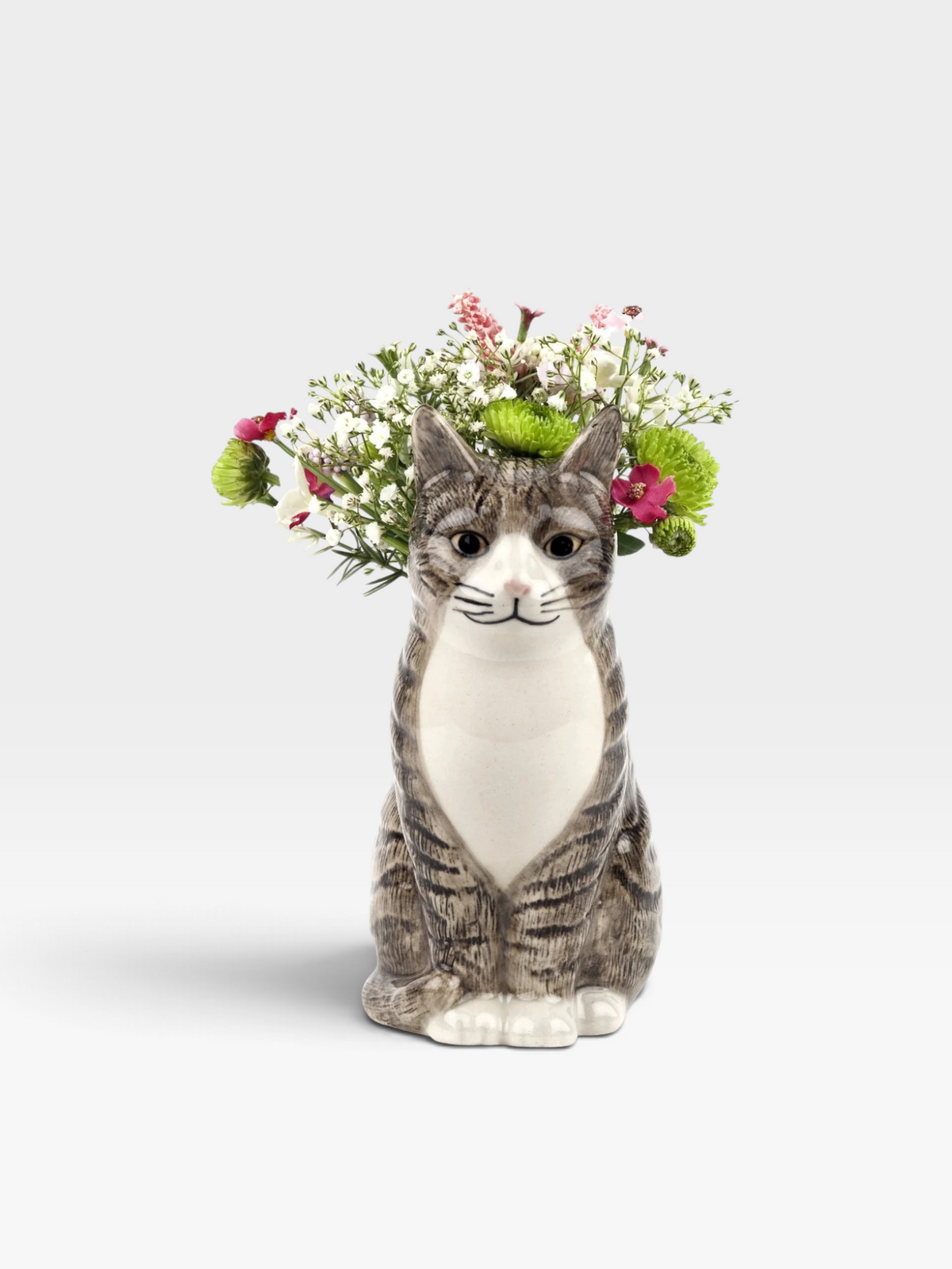 A ceramic cat-shaped bud vase with a gray and white tabby cat design, holding a small arrangement of pink and green flowers, set against a light gray background.