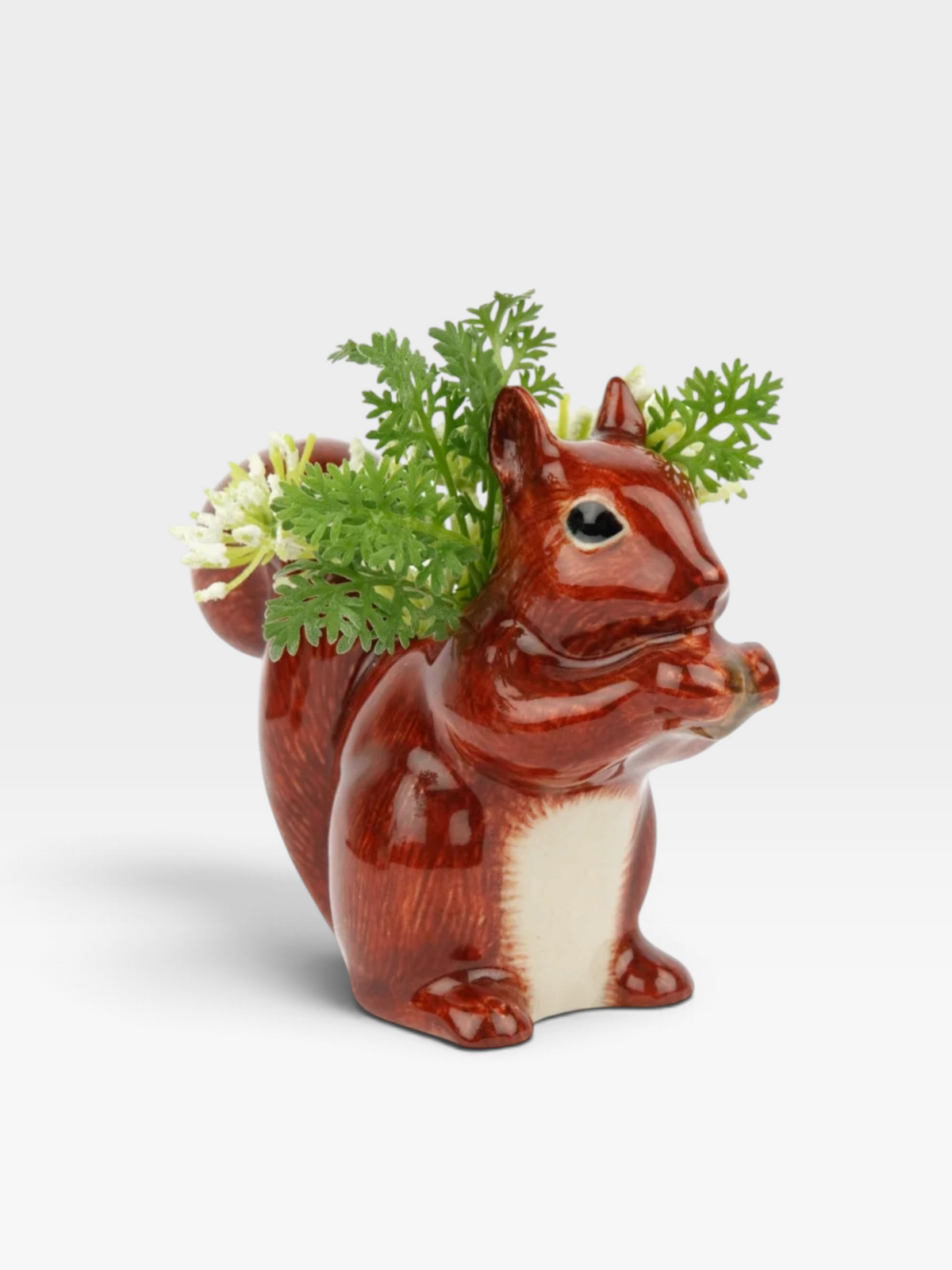 A glossy ceramic squirrel bud vase in reddish-brown and cream glaze, holding small green and white flowers, photographed on a clean light gray background.