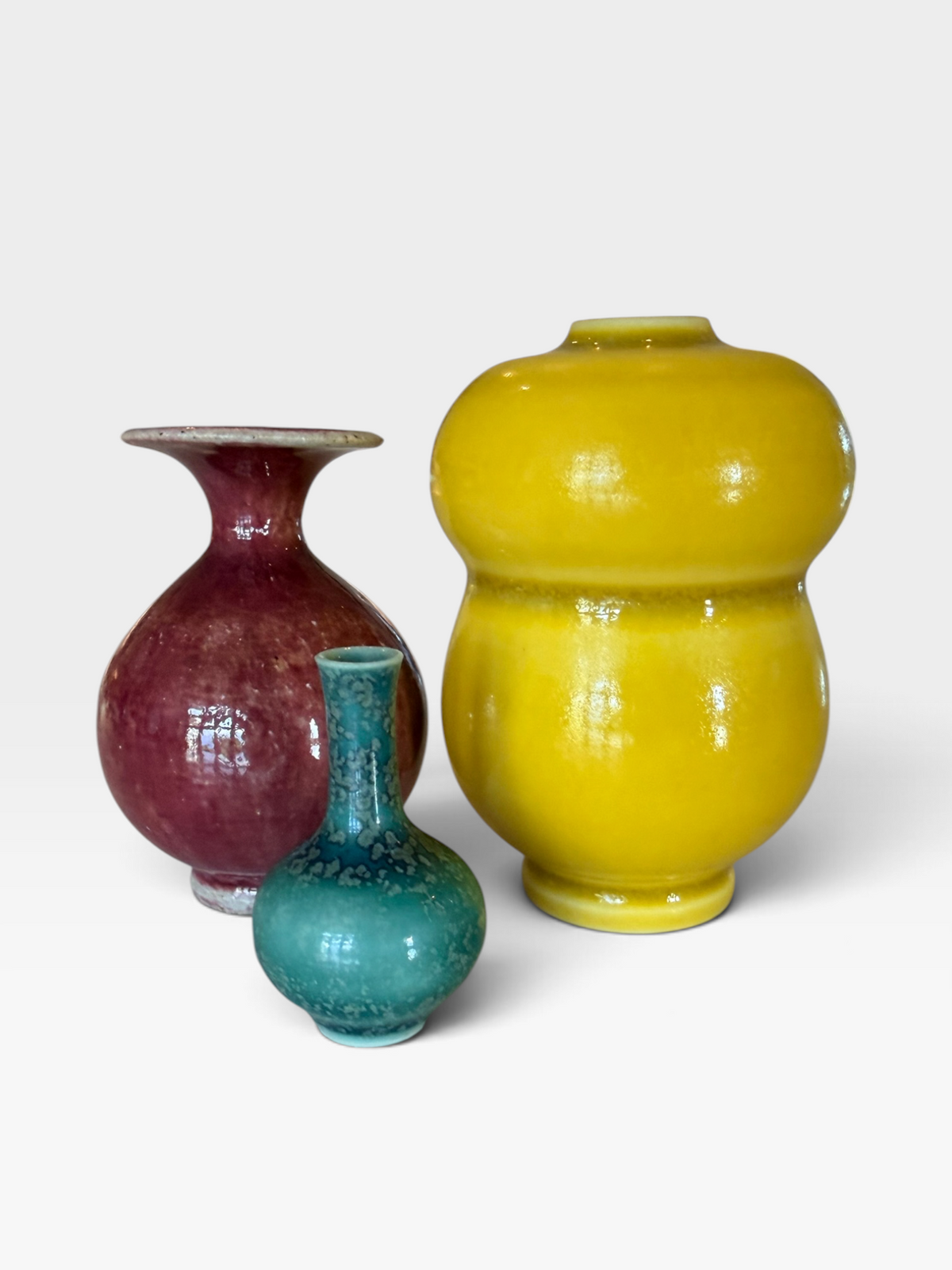 Three glazed ceramic vases in yellow, burgundy, and teal, arranged on a light gray background.