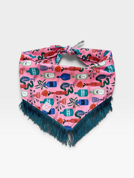 Pink dog bandana with colorful bottle pattern and teal-colored fringe on a textured white background