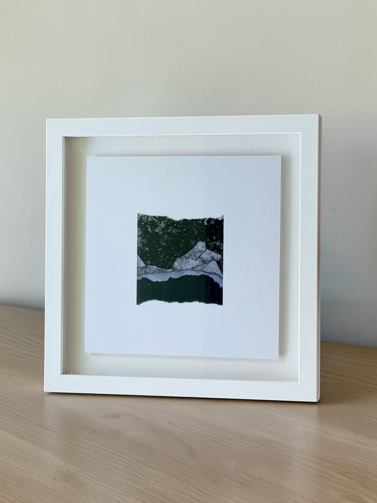 A white square shadow box frame containing a small abstract landscape art print, displayed on a light wood surface against a neutral beige wall.