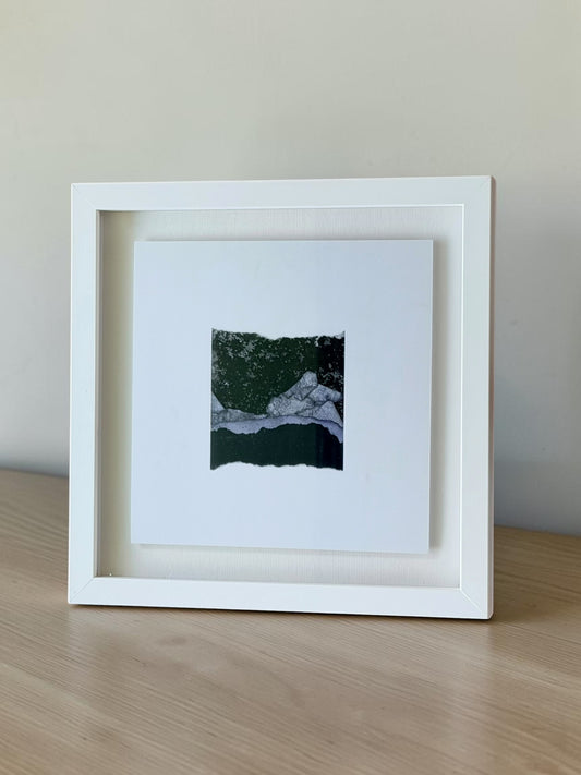 A white square shadow box frame containing a small abstract landscape art print, displayed on a light wood surface against a neutral beige wall.