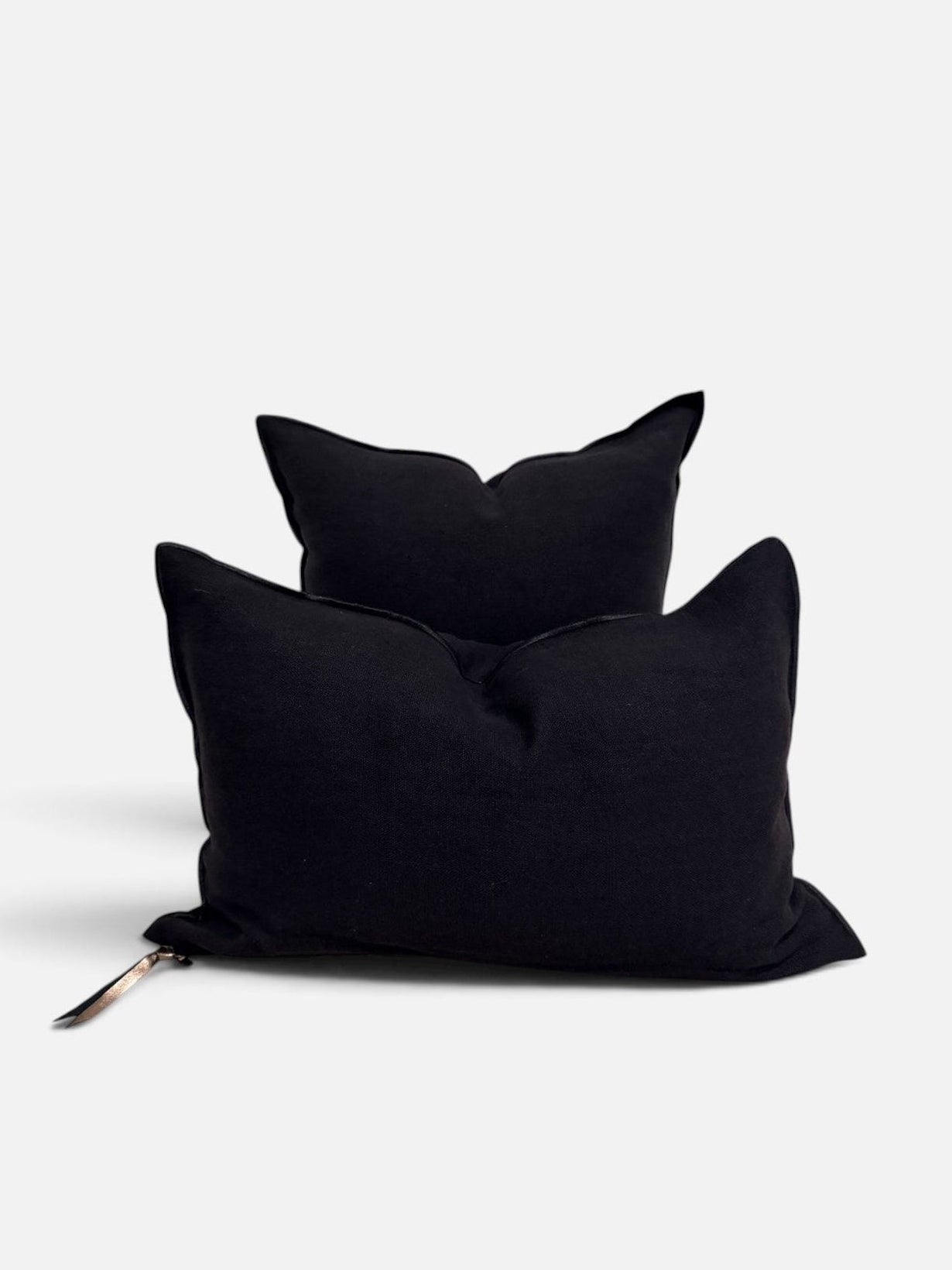 Two black rectangular throw pillows stacked together on a light gray background, with a small metallic zipper pull visible on the lower pillow.