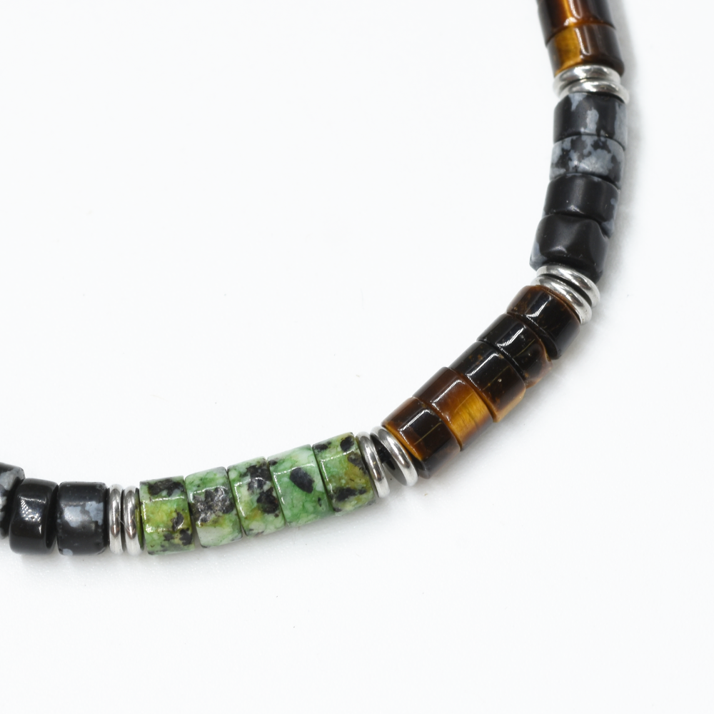 A close-up of a beaded necklace featuring faceted green and black beads, polished brown tiger's eye beads, and silver spacer rings on a white background.