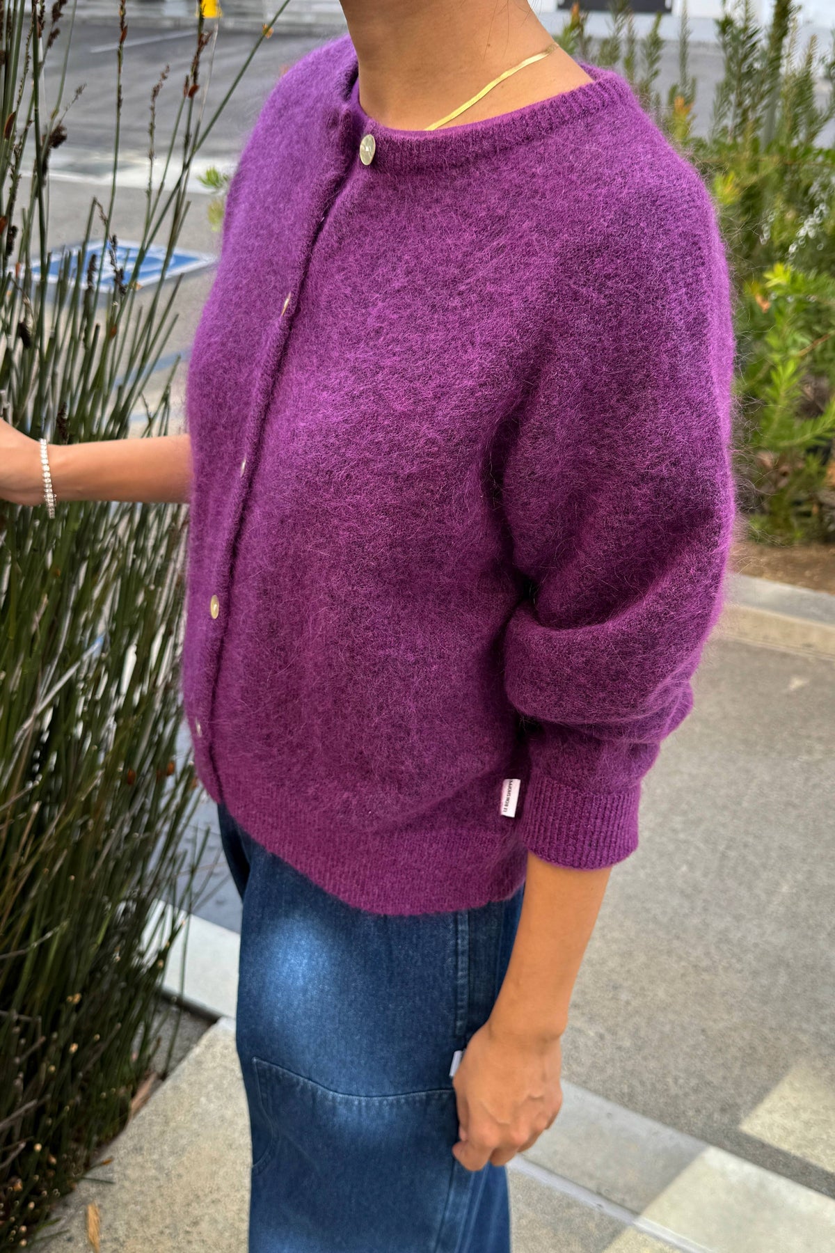 A person wearing a fuzzy purple knit cardigan with a round neckline, front buttons, and ribbed cuffs, paired with a blue denim skirt outdoors.