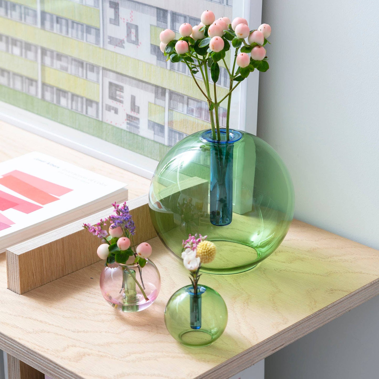 A collection of bubble glass vases in green and blue colors, with one large green vase, a small blue vase, and two smaller pink vases, arranged on a table with a window in the background.