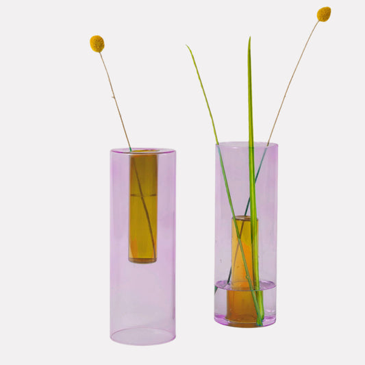 Two reversible glass vases, one with a peach color and the other with a lilac color, each containing a single green stem. The vases are designed to hold multiple stems when flipped.
