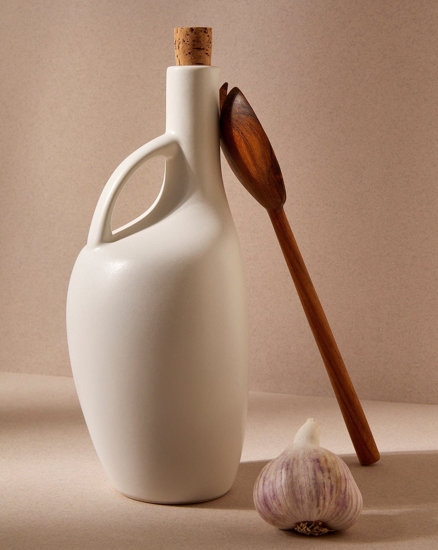 A white ceramic olive oil with cork stopper sits on a beige surface with a head of garlic sitting beside it and a tall wooden spoon standing upright leaning against it