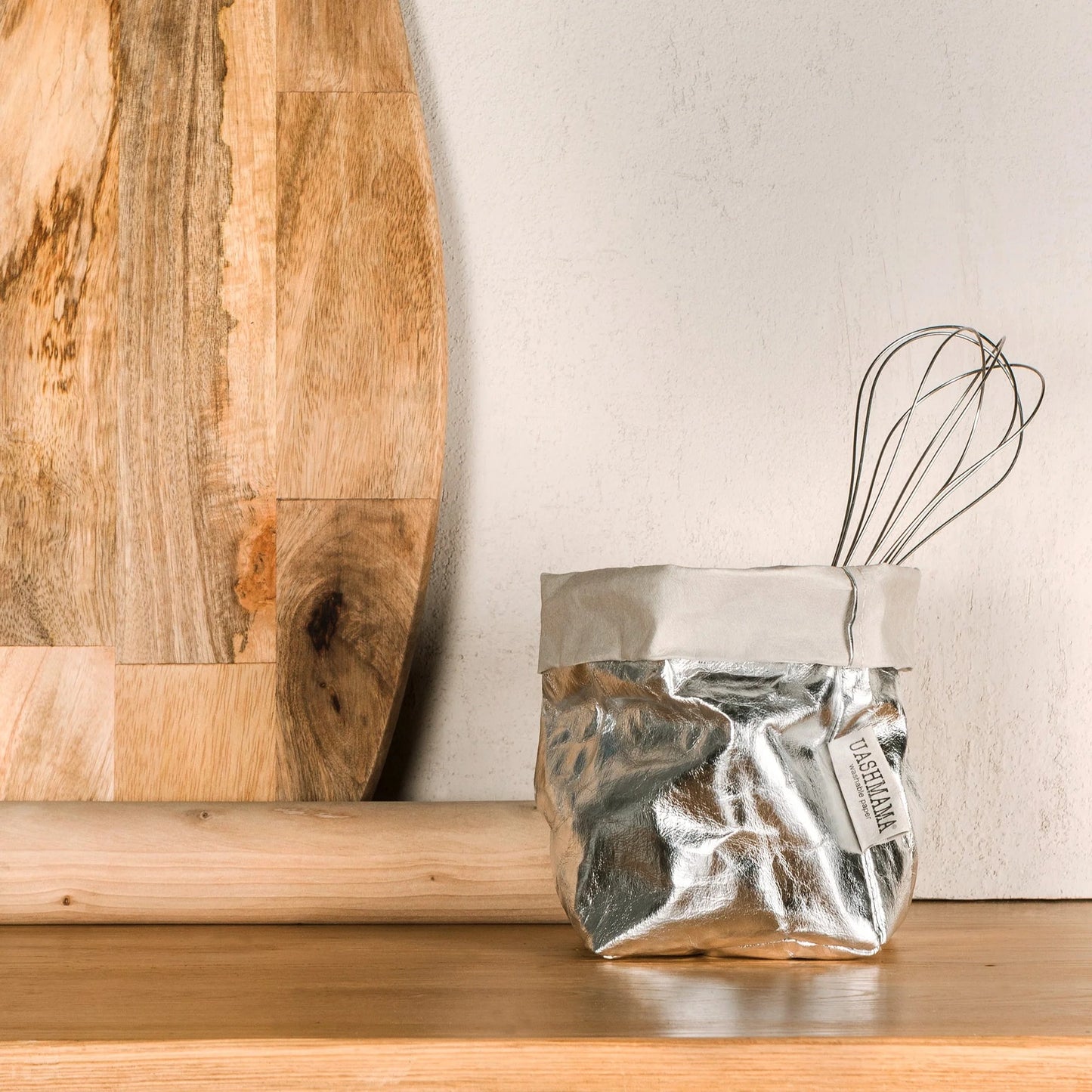 Silver container with a whisk on a wooden surface next to a wooden cutting board.