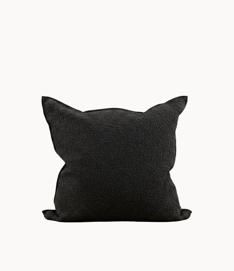 A black textured throw pillow with cat-ear points, resting on a white surface against a plain off-white background.