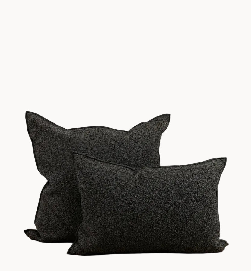 Two black textured throw pillows, one standing upright and one leaning flat, displayed on a light wood bench against a plain off-white wall.