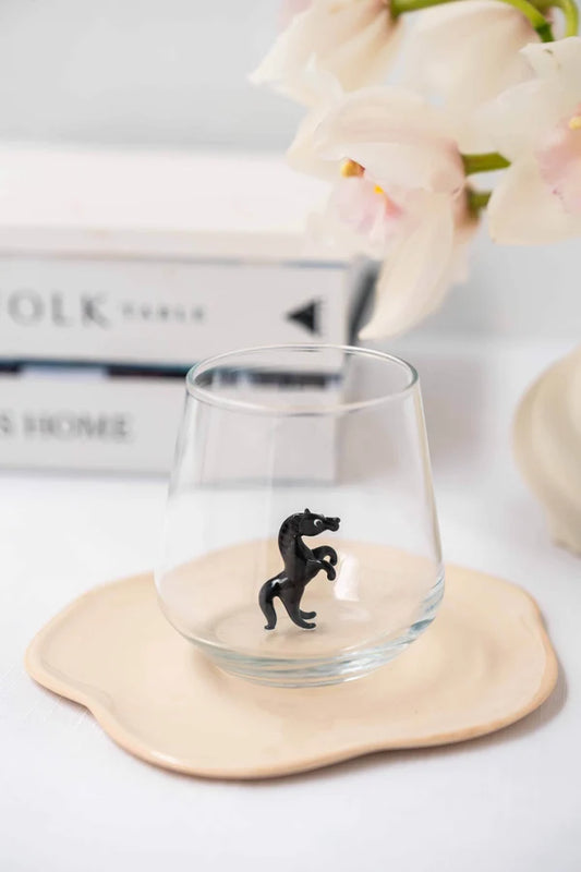 A clear stemless glass tumbler with a small black horse figurine inside, resting on a beige ceramic coaster. The background is softly blurred with white flowers and a book.