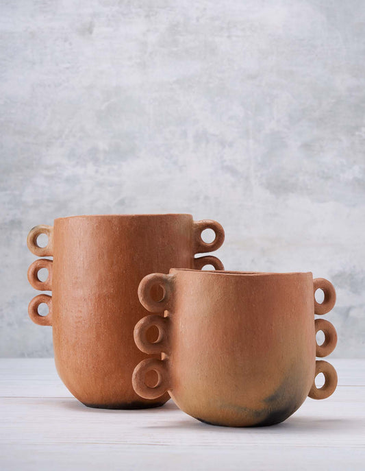 A small terracotta pot sits in front of a larger terracotta pot, both have ring details on the sides and are set on a white surface against a light grey background