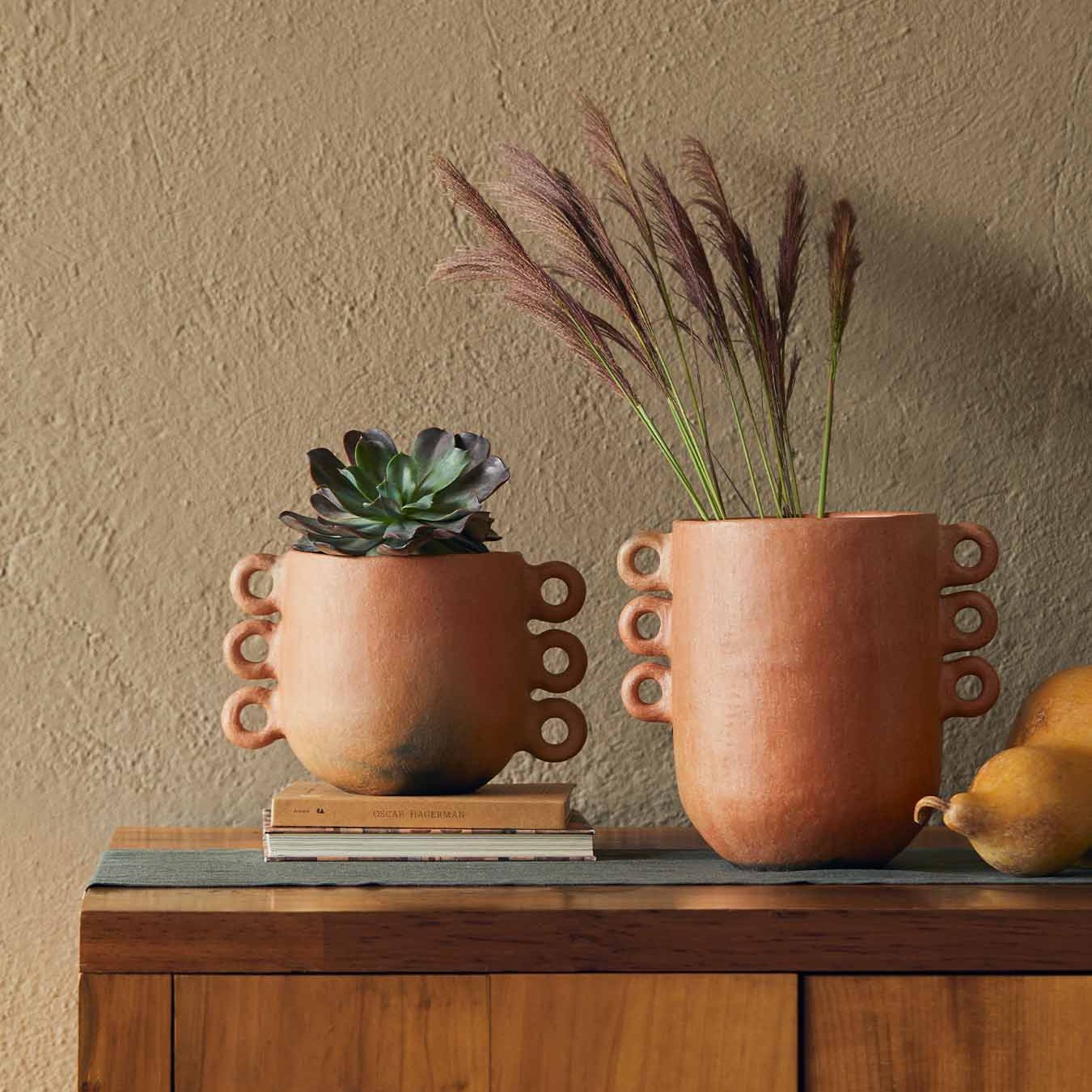 Two terracotta pots with decorative rings on the sides sit on a table, one large and one small, used as planters for a succulent and some dried florals