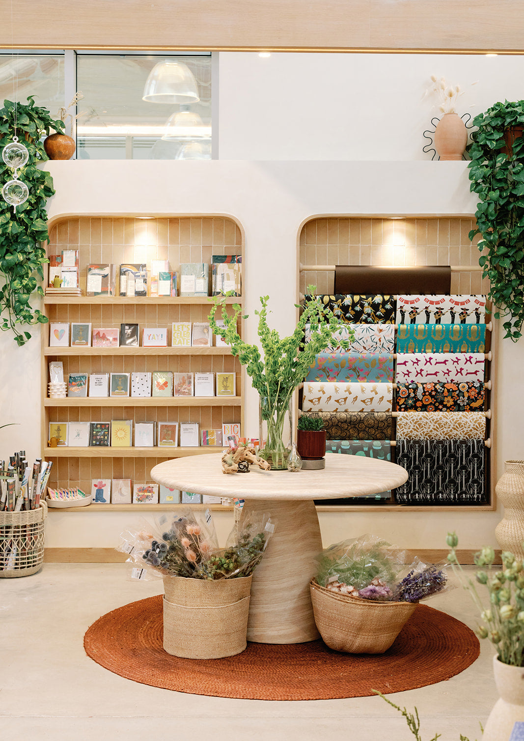 Interior of a store with shelves displaying books, cards, wrapping paper, dried floral bouquets and decorative items, featuring a round table with plants.