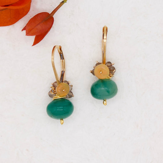 Gold dangle earrings with green, topaz, and yellow stones sit on a white surface with red flowers visible in the top left corner
